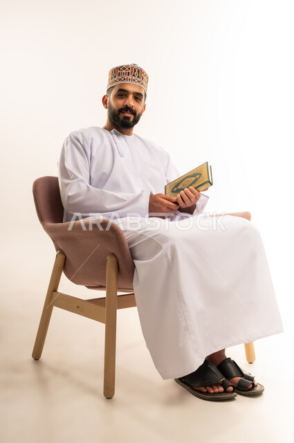 الشعور بالخشوع والطمأنينة، تلاوة وقراءة القرآن الكريم وتدبر معانيه، بورتريه  لرجل عربي خليجي عماني يرتدي الدشداشة والكمة يحمل بيده المصحف الشريف،الإكثار من ذكر الله تعالى، خلفية بيضاء .