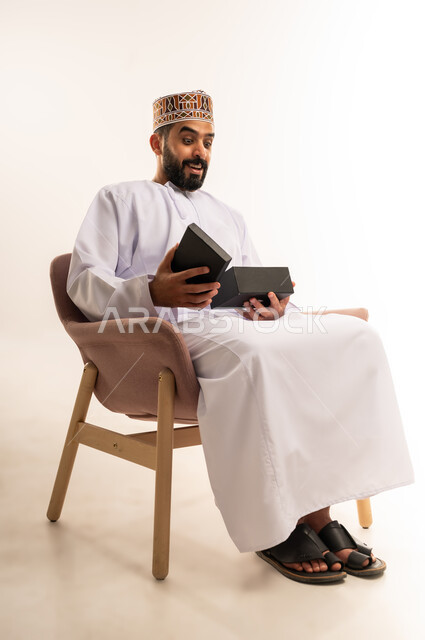 إيماءات الذهول والمفاجأة ، مفهوم الشكر والامتنان، تقديم الهدايا بالاعياد والمناسبات الخاصة، التعبير عن الحب والتقدير والاحترام، بورتريه لرجل عربي خليجي عماني يرتدي الدشداشة والعمامة يجلس على كرسى مريح يقوم بفتح صندوق الهدية بتعابيرالسرور والسعادة، خلفية بيضاء .