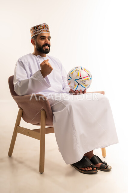 Enjoying watching your favorite team, having a good time following the match, portrait of a young Gulf Arab Omani man wearing a dishdasha and a kummah sitting on a comfortable chair holding a football with an expression of enthusiasm, raising his fist with gestures of victory and triumph, matches and sports, white background.