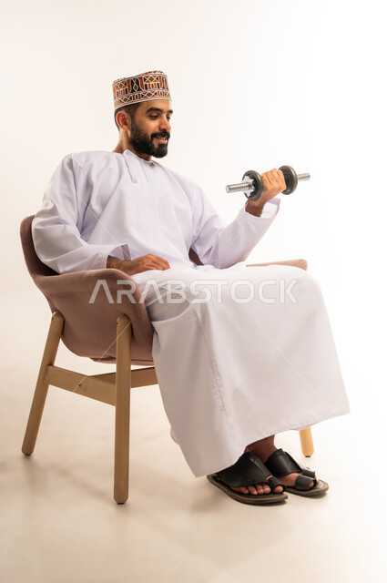 نشاط بدني للمحافظة على اللياقة الجسدية، رفع الأوزان الحديدية الثقيلة، بناء عضلات الجسم وتقويتها، بورتريه لرجل عربي خليجي عماني يرتدي الدشداشة والكمة يجلس على الاريكة يحمل بيده الأثقال، خلفية بيضاء.