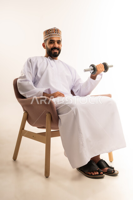 بناء عضلات الجسم وتقويتها، نشاط بدني للمحافظة على اللياقة الجسدية، رفع الأوزان الحديدية الثقيلة، بورتريه لرجل عربي خليجي عماني يرتدي الدشداشة والكمة يجلس على الاريكة يحمل بيده الأثقال، خلفية بيضاء.