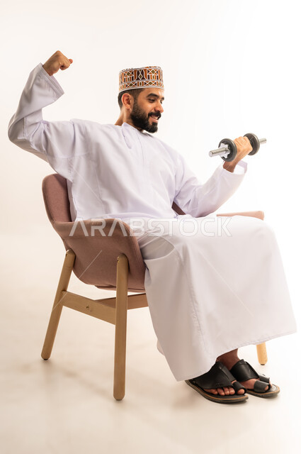 رفع الأوزان الحديدية الثقيلة، بناء عضلات الجسم وتقويتها، نشاط بدني للمحافظة على اللياقة الجسدية، بورتريه لرجل عربي خليجي عماني يرتدي الدشداشة والكمة يجلس على الاريكة يحمل بيده الأثقال، خلفية بيضاء.