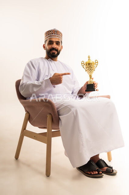 قضاء وقت ممتع بمتابعة المباراة، المباريات والألعاب الرياضية، الاستمتاع بمشاهدة الفريق المفضل،بورتريه لرجل عربي خليجي عماني يرتدي الدشداشة والكمة يجلس على أريكة مريحة يمسك الكأس ويشير اليه بتعابير الحماس، خلفية بيضاء .