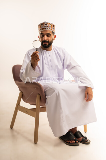 مفهوم الفحص والاستكشاف، توضيح الصورة والرؤية عن قرب، بورتريه لشاب عربي خليجي عماني يرتدي الكمة و الدشداشة يجلس على كرسي مريح يحمل بيده عدسة مكبرة ينظر من خلالها بإيماءات التركيز والتمعن، خلفية بيضاء .