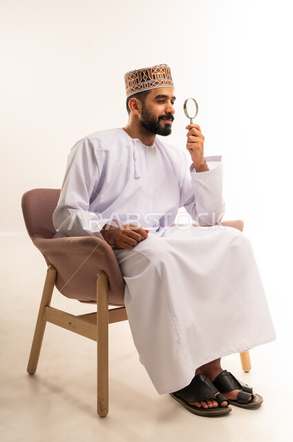 أدوات بصرية للتدقيق والتركيز، مفهوم الفحص والاستكشاف، توضيح الصورة والرؤية عن قرب، بورتريه لشاب عربي خليجي عماني يرتدي الكمة و الدشداشة يجلس على كرسي مريح يحمل بيده عدسة مكبرة ينظر من خلالها بإيماءات التركيز والتمعن، خلفية بيضاء .