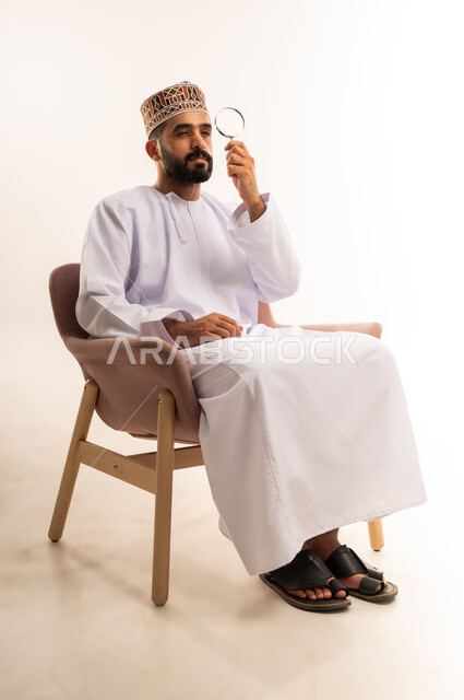 توضيح الصورة والرؤية عن قرب، بورتريه لشاب عربي خليجي عماني يرتدي الكمة و الدشداشة يجلس على كرسي مريح يحمل بيده عدسة مكبرة ينظر من خلالها بإيماءات التركيز والتمعن، مفهوم الفحص والاستكشاف، خلفية بيضاء .