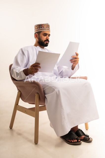 أدوات تدوين المهام اليومية، استخدام الورق الأبيض في تسجيل الملاحظات والمعلومات المهمة, بورتريه لرجل عربي خليجي عماني يرتدي الدشداشة والكمة يمسك بيدة الاوراق، بإيماءات التركيز والتمعن، صورة الجسم بالطول الكامل ،خلفية بيضاء .