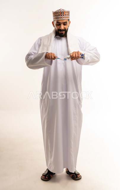 استخدام منتجات محلية وطنية، المحافظة على ابتسامة بيضاء، تجنب التسوس وأمراض اللثة، مفهوم العناية والإهتمام بصحة الأسنان، روتين نظافة الفم اليومية، بورتريه لرجل عربي خليجي عماني يرتدي الدشداشة والكمة يقوم بتنظيف أسنانه باستخدام الفرشاة والمعجون، صورة الجسم بالطول الكامل ، خلفية بيضاء .