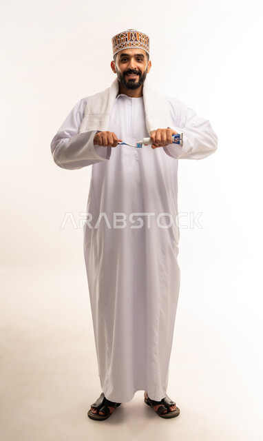 Avoiding cavities and gum diseases, the concept of dental care and attention, daily oral hygiene routine, portrait of an Arab Gulf Omani man wearing a dishdasha and a kuma cleaning his teeth using a toothbrush and toothpaste, full body image, white background.