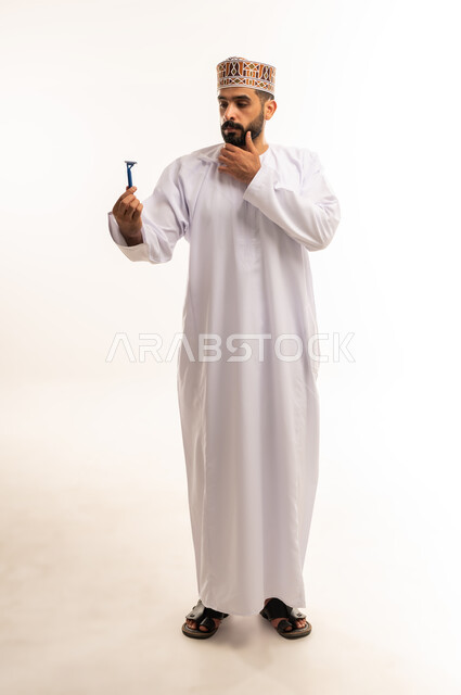 روتين العناية الشخصية ، المحافظة على الأناقة والنظافة الشخصية، القيام بإزالة شعر الوجه ، الاهتمام بالمظهر الخارجي، بورتريه لرجل عربي خليجي عماني يرتدي الدشداشة والكمة يمسك بيده شفرة الحلاقة الرجالية وينظر إليها ، صورة الجسم بالطول الكامل، خلفية بيضاء .