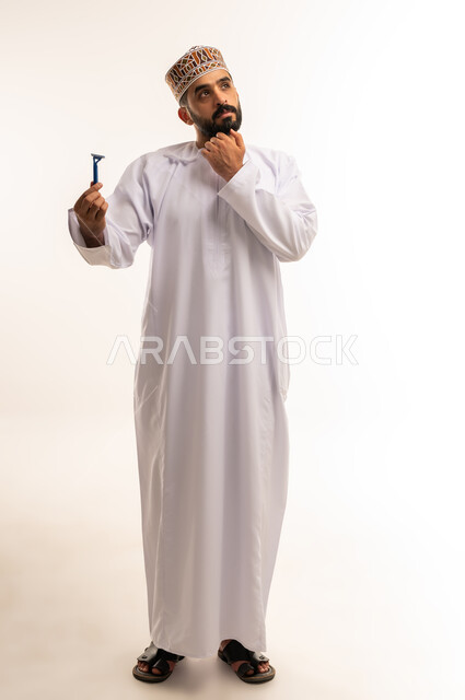 روتين الحلاقة الرجالية ، المحافظة على الأناقة والنظافة الشخصية، القيام بإزالة شعر الوجه ، الاهتمام بالمظهر الخارجي، بورتريه لرجل عربي خليجي عماني يرتدي الدشداشة والكمة يمسك بيده الشفرة وينظر إليها بايماءات الاستغراب ، صورة الجسم بالطول الكامل، خلفية بيضاء .
