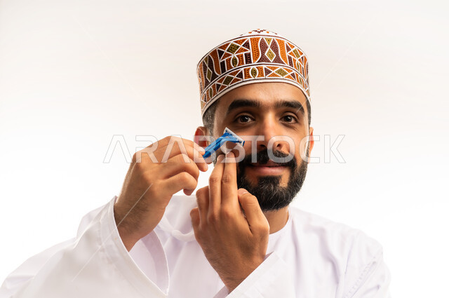 وعي الرجل العربي بالعناية الشخصية ، روتين الحلاقة الرجالية ، القيام بإزالة شعر الوجه ، الاهتمام بالاناقة والمظهر الخارجي، بورتريه مقرب لرجل عربي خليجي عماني يرتدي الدشداشة والكمة يمسك بيده الشفرة ، خلفية بيضاء .