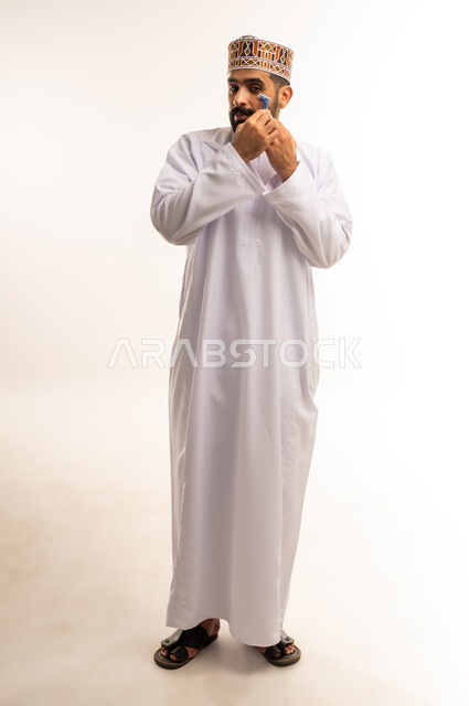 الاهتمام بالمظهر الخارجي، النظر الى الكاميرا بإيماءات الفرح والسرور، المحافظة على الأناقة والنظافة الشخصية، بورتريه لرجل عربي خليجي عماني يرتدي الدشداشة والكمة ، القيام بإزالة شعر الوجه باستخدام شفرة الحلاقة الرجالية، الجسم بالطول الكامل، خلفية بيضاء .