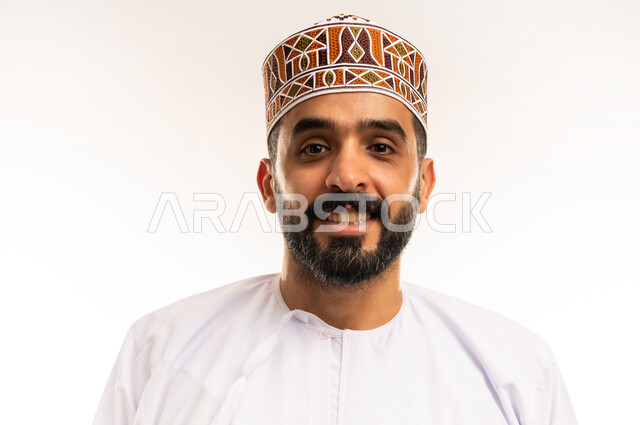 مفهوم الرجولة والثقة بالنفس، التقاط الصور الشخصية للمناسبات العامه، الاهتمام بالأناقة والمظهر الخارجي,  بورتريه مقرب لرجل عربي خليجي عماني مبتسم يرتدي الدشداشة و الكمه ينظر الى الكاميرا بإيماءات السرور والسعادة،  خلفية بيضاء .