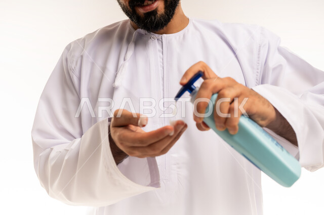 استخدام الكريمات ومكونات التجميل الشبابية، مفهوم العناية والاهتمام بنضارة البشرة وتحسينها، صورة مقربة ليد رجل عربي خليجي عماني يرتدي الدشداشة والكمة يحمل بيده عبوة لكريم مرطب، خلفية بيضاء.