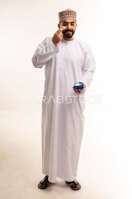 استخدام مكونات تجميلية شبابية، ترطيب وتنظيف وتفتيح الجلد، بورتريه لرجل عربي خليجي عماني يرتدي الدشداشة والكمة يضع كريم المرطب على الوجه، مفهوم العناية والإهتمام بنضارة وتحسين البشرة، الجسم بالطول الكامل، خلفية بيضاء.