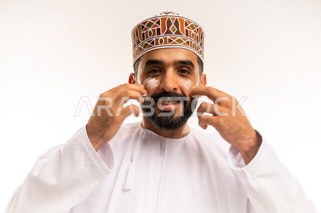 روتين العناية بالشرة ، ترطيب وتنظيف وتفتيح الجلد، مفهوم الإهتمام بنضارة وتحسين البشرة، استخدام مكونات تجميلية شبابية، بورتريه مقرب لرجل عربي خليجي عماني يرتدي الدشداشة والكمة يضع كريم المرطب على الوجه، خلفية بيضاء.