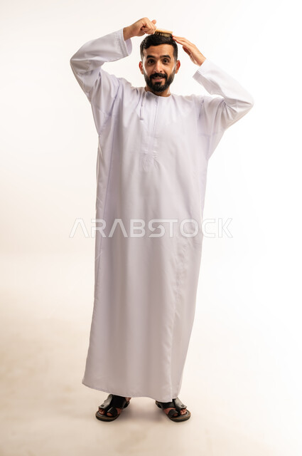 المحافظة على شعر صحي وأنيق، مفهوم الاهتمام بالجمال والأناقة والمظهر الخارجي، العناية بالنظافة الشخصية, ترتيب وتمشيط الشعر، بورتريه لرجل عربي خليجي عماني يرتدى الدشداشة ، يقوم بتصفيف شعره باستخدام فرشاة الشعر ( المشط ) بتعابير وإيماءات تدل على السعادة والسرور، صورة الجسم بالطول الكامل ،خلفية بيضاء .