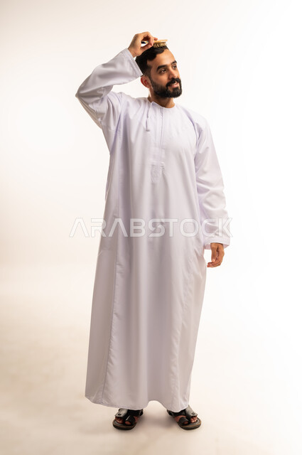 مفهوم الاهتمام بالجمال والأناقة والمظهر الخارجي، العناية بالنظافة الشخصية, ترتيب وتمشيط الشعر، بورتريه لرجل عربي خليجي عماني يرتدى الدشداشة ، يقوم بتصفيف شعره باستخدام فرشاة الشعر ( المشط ) بتعابير وإيماءات تدل على السعادة والسرور، صورة الجسم بالطول الكامل ،خلفية بيضاء .