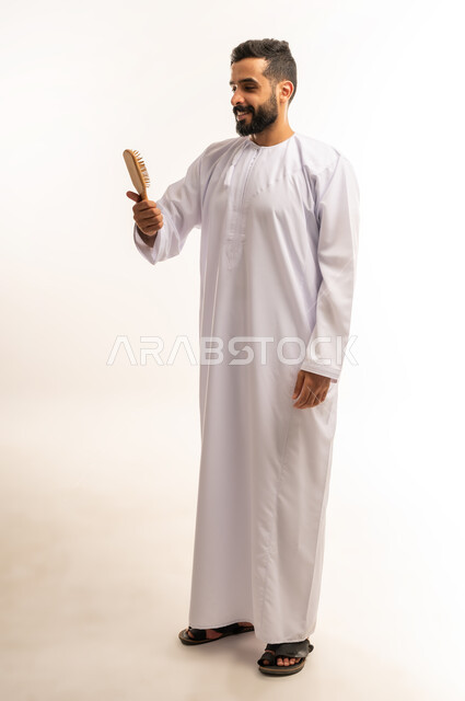 Personal hygiene, hair styling and combing, the concept of caring for beauty, elegance and outward appearance, a portrait of an Arab Gulf Omani man wearing a dishdasha, styling his hair using a hairbrush (comb) with expressions and gestures indicating happiness and pleasure, a full-length body image, a white background.