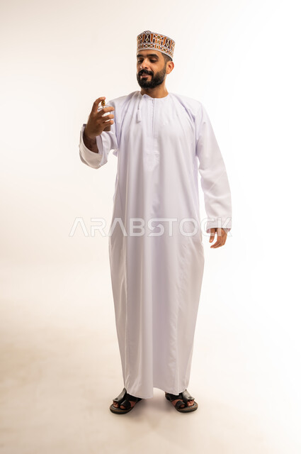 الاستعداد للذهاب والخروج من المنزل, التسويق للعطور الخليجية العمانية, استخدام الزيوت العطرية الطبيعية, بورتريه لرجل عربي خليجي عماني مبتسم يرتدي الدشداشه و الكمه يقوم برش العطر بتعابير السرور, صورة الجسم بالطول الكامل, خلفية بيضاء .