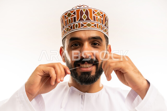 المحافظة على ابتسامة ناصعه البياض ، التأكد من نظافة الأسنان بعد تناول الطعام ، تنظيف الأسنان بالخيط السني ، الاهتمام بالأناقة والمظهر الخارجي، مفهوم العناية بصحة الفم، بورتريه مقرب لرجل عربي خليجي عماني يرتدى الدشداشة والكمة ، خلفية بيضاء .