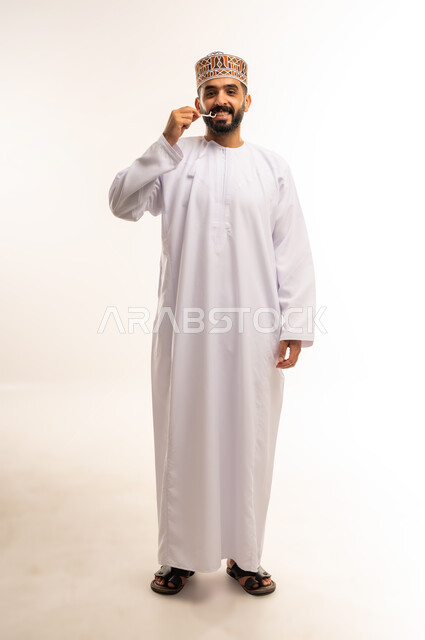 مفهوم العناية بصحة الفم، التأكد من نظافة الأسنان بعد تناول الطعام، الاهتمام بالأناقة والمظهر الخارجي، المحافظة على ابتسامة بيضاء، بورتريه  لرجل عربي خليجي عماني يقوم بتنظيف أسنانه باستخدام الخيط السني، صورة الجسم بالكامل، خلفية بيضاء .