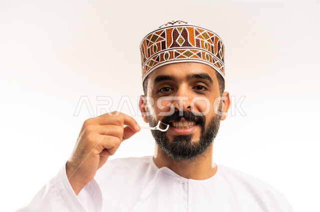 معدات طب الأسنان ، استخدام الخيط السني ، الاهتمام بالأناقة والمظهر الخارجي، المحافظة على ابتسامة بيضاء، مفهوم العناية بصحة الفم، التأكد من نظافة الأسنان بعد تناول الطعام، بورتريه مقرب لرجل عربي خليجي عماني يرتدى الدشداشة والكمة ، يقوم بتنظيف أسنانه ، خلفية بيضاء .