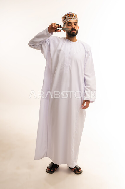 استخدام أدوات و معدات الحلاقة, صالون حلاقة تجميلي رجالي، العناية بشعر صحي ونظيف, ، بورتريه لرجل عربي خليجي عمانى يرتدى الدشداشة والكمة ، يحمل بيده المقص ، قص و تصفيف الشعر ، مفهوم الإهتمام بالجمال والمظهر الخارجي ، الجسم بالطول الكامل ،خلفية بيضاء .