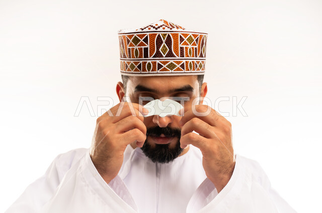 التخلص من الرؤوس السوداء والجلد الميت، مفهوم العناية والاهتمام بنضارة وتحسين البشرة، استخدام ماسكات تنظيف المسام ومكونات تجميلية، بورتريه مقرب لرجل عربي خليجي عماني يرتدي الدشداشة والكمة يضع لصقات الأنف ، مكونات تجميلية شبابية، خلفية بيضاء .