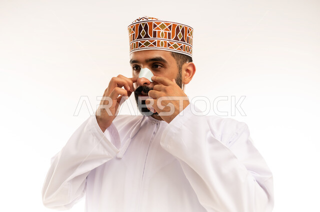 استخدام ماسكات تنظيف المسام ومكونات تجميلية، بورتريه مقرب لرجل عربي خليجي عماني يرتدي الدشداشة والكمة يضع لصقات الأنف للتخلص من الرؤوس السوداء والجلد الميت، مفهوم العناية والاهتمام بنضارة وتحسين البشرة، خلفية بيضاء .