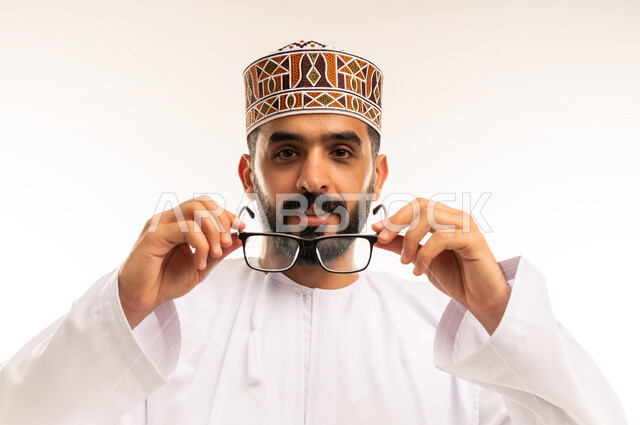 مفهوم الضبابية وعدم وضوح الرؤية ، بورتريه مقرب لرجل عربي خليجي عماني يرتدي الدشداشة والكمة ,يلبس النظارات الطبية وينظر الى الكاميرا بإيماءات السعادة ، العدسات الخاصة بضعف النظر ، استخدام البصريات لتوضيح الرؤية، خلفية بيضاء .