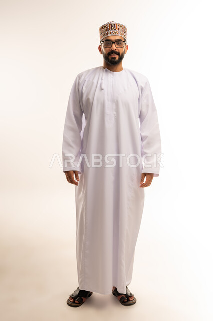 الرؤية من خلال العدسات الطبية, إيماءات السعادة و السرور بوضوح الصورة، بورتريه لرجل عربي خليجي عماني يرتدي الدشداشة والكمة ,يلبس النظارات الطبية وينظر الى الكاميرا ، استخدام البصريات لتوضيح الرؤية، الجسم بالطول الكامل ،خلفية بيضاء .