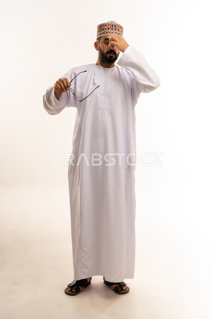 النسيان ومحاولة تذكر شيء ما, الاحساس بالشقيقة والصداع النصفي ، بورتريه لرجل عربي خليجي عماني يرتدي الدشداشة والكمة ،يضع يده على عينه ويغمضها ، الرؤية من خلال العدسات الطبية, استخدام البصريات لتوضيح الرؤية، الجسم بالطول الكامل ،خلفية بيضاء .