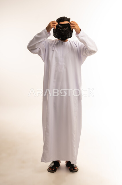 مقهوم النقاء بماسك الفحم الفعّال ، الاستمتاع بلحظات العناية الذاتية ، استخدام مكونات تجميلية، ماسك تنظيف وتقشير البشرة، بورتريه لشاب عربي عماني خليجي يرتدي الدشداشة يقوم بوضع ماسك على وجهه للتخلص من الرؤوس السوداء ، الجسم بالطول الكامل، خلفية بيضاء .