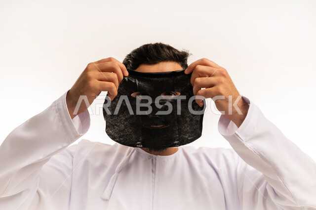 التنفس النقى للبشرة ، الاهتمام بالمظهر الخارجي، الاستمتاع بلحظات العناية الذاتية ، استخدام مكونات تجميلية، ماسك تنظيف وتقشير البشرة، بورتريه مقرب لشاب عربي عماني خليجي يرتدي الدشداشة يقوم بوضع ماسك على وجهه للتخلص من الرؤوس السوداء ، خلفية بيضاء .