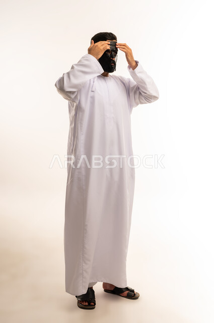 استخدام قناع الفحم الاسود ، مفهوم العناية والاهتمام بالمظهر الخارجي، استخدام مكونات تجميلية، ماسك تنظيف وتقشير البشرة، بورتريه لشاب عربي عماني خليجي يرتدي الدشداشة يقوم بوضع ماسك على وجهه للتخلص من الرؤوس السوداء، الجسم بالطول الكامل، خلفية بيضاء .