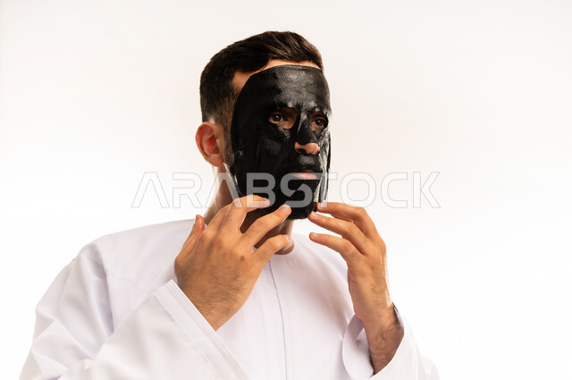 مفهوم العناية والاهتمام بالمظهر الخارجي، استخدام مكونات تجميلية، ماسك تنظيف وتقشير البشرة، بورتريه مقرب لشاب عربي عماني خليجي يرتدي الدشداشة يقوم بوضع قناع الفحم الاسود على وجهه للتخلص من الرؤوس السوداء، خلفية بيضاء .