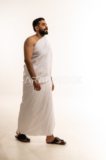 Getting closer to God by performing the Hajj rituals, the fifth pillar of Islam, performing the Hajj pilgrimage, side portrait of a young Omani Arab Gulf man wearing Ihram clothes and a mask looking at the camera, visiting the Holy House of God in Mecca, full body photo, white background.