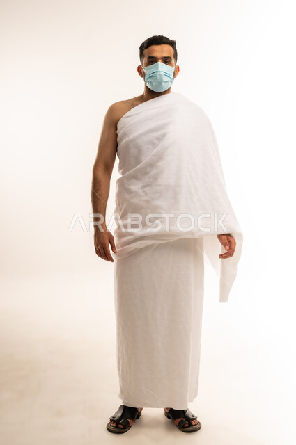Visiting the Holy House of Allah in Mecca, performing the Hajj pilgrimage, portrait of a young Omani Arab Gulf man wearing Ihram clothes and a mask looking at the camera, full body photo, white background.