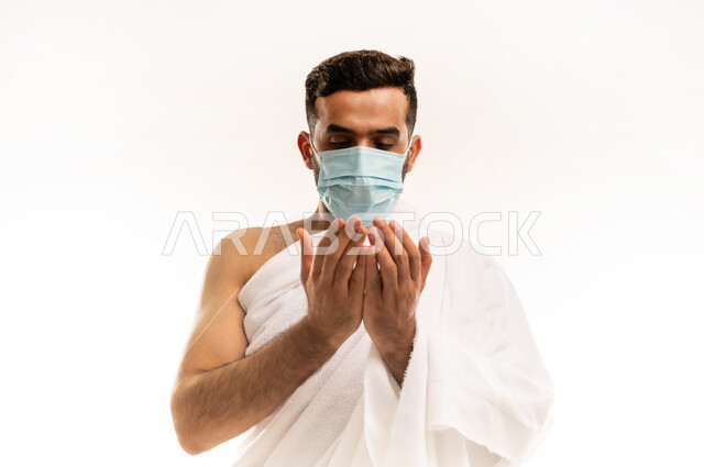 التقرب الى الله بالأعمال الصالحة, أدعية وأذكار اسلامية ، الركن الخامس من أركان الاسلام ، مفهوم التضرع والعبادة , بورتريه مقرب لرجل عربي عماني خليجي يرتدي ملابس الإحرام  والكمامة ، يرفع يديه للأعلى ويدعى بخشوع ، الاجتهاد بالأدعية والطاعات ، الجسم بالطول الكامل، خلفية بيضاء .