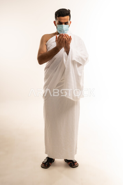 مفهوم العبادة والتقرب إلى الله تعالى, الاجتهاد بالأدعية والطاعات والأذكار, بورتريه من الجانب لرجل عربي عماني خليجي يرتدي ملابس الإحرام  والكمامة ، يرفع يديه للأعلى بتعابير الدعاء ، الجسم بالطول الكامل، خلفية بيضاء .