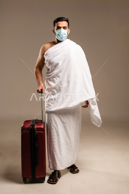 Preparing to head out and set off to visit the Sacred House of God, perform the Hajj and Umrah pilgrimages, worship, obedience and drawing closer to God, portrait of an Omani Arab Gulf man wearing Ihram clothes, holding a travel bag and wearing a mask, full body, white background.