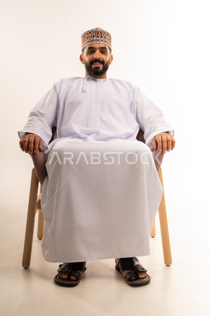 الاسترخاء والاستمتاع بالأجواء الهادئة، مفهوم الاناقة والاهتمام بالمظهر الخارجي, بورتريه لرجل عربي خليجي عماني يرتدي الدشداشه والكمه يجلس على كرسي مريح ينظر الى الكاميرا بايماءات السعادة والسرور، خلفية بيضاء .