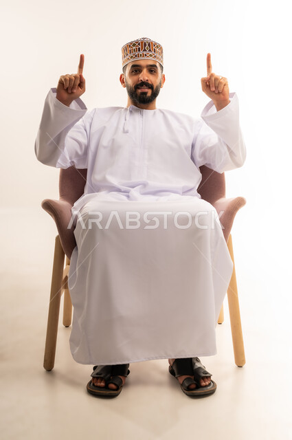 تعبيرات البشاشة والسرور , رفع اليدين إلى الأعلى والنظر إلى الكاميرا, ايماءات التأكيد وتحديد شيء معين ، بورتريه لشاب عربي خليجي عماني يرتدي الدشداشة والكمه يجلس على كرسى مريح ويشير بإصابع السبابة إلى شيء ما, خلفية بيضاء .