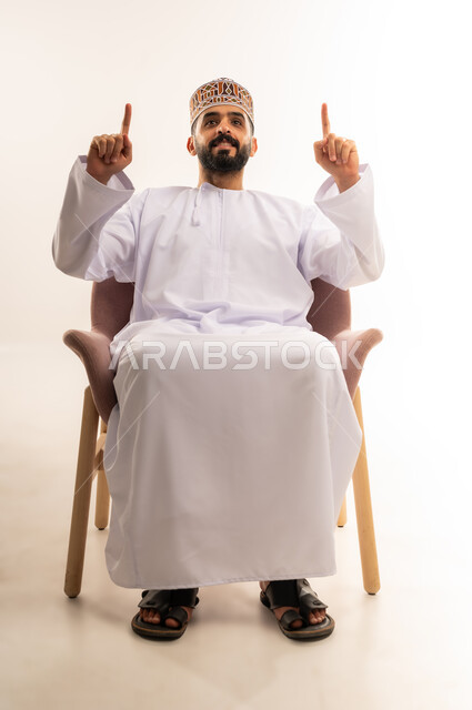 ايماءات التأكيد وتحديد شيء معين ، رفع اليدين إلى الأعلى والنظر إلى الكاميرا,تعبيرات البشاشة والسرور , بورتريه لشاب عربي خليجي عماني يرتدي الدشداشة والكمه يجلس على كرسى مريح ويشير بإصابع السبابة إلى شيء ما, خلفية بيضاء .