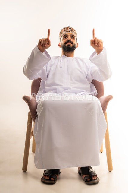رفع اليدين إلى الأعلى والنظر إلى الكاميرا, البشاشة والسرور وتحديد شيء معين, بورتريه لشاب عربي خليجي عماني يرتدي الدشداشة والكمه يجلس على كرسى مريح ويشير بإصابع السبابة إلى شيء ما, خلفية بيضاء .