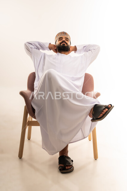 مفهوم الاسترخاء والاستمتاع بالأجواء الهادئة، بورتريه لرجل عربي خليجي عماني يرتدي الدشداشه والكمة يجلس على كرسي مريح ويضع يديه تحت رأسه، الشعور بالتعب والحاجة إلى النوم، ايماءات تدل على الراحة، خلفية بيضاء .