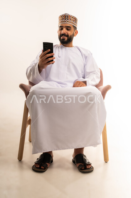 التواصل مع الأصدقاء عبر مواقع التواصل الاجتماعي، متابعة الاحداث العالمية باستخدام الهاتف المحمول، استخدام الأجهزة التقنية الحديثة ، بورتريه لشاب عربي خليجي عماني يرتدي الدشداشة والكمة ويمسك بيده الجوال ، خلفية بيضاء .