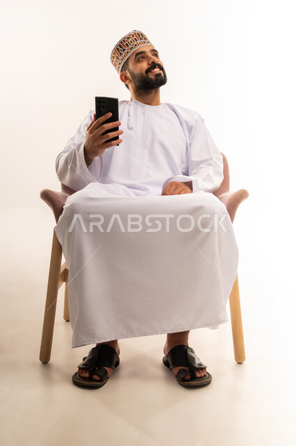 تصفح برامج وتطبيقات الجوال ، استخدام الأجهزة التقنية الحديثة ،التواصل مع الأصدقاء عبر مواقع التواصل الاجتماعي، متابعة الاحداث العالمية باستخدام الهاتف المحمول ، بورتريه لشاب عربي خليجي عماني يرتدي الدشداشة والكمة ويمسك بيده الجوال ، خلفية بيضاء .